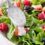 A spoon drizzles creamy poppy seed dressing over a salad with romaine lettuce, strawberries, raspberries, and blackberries.