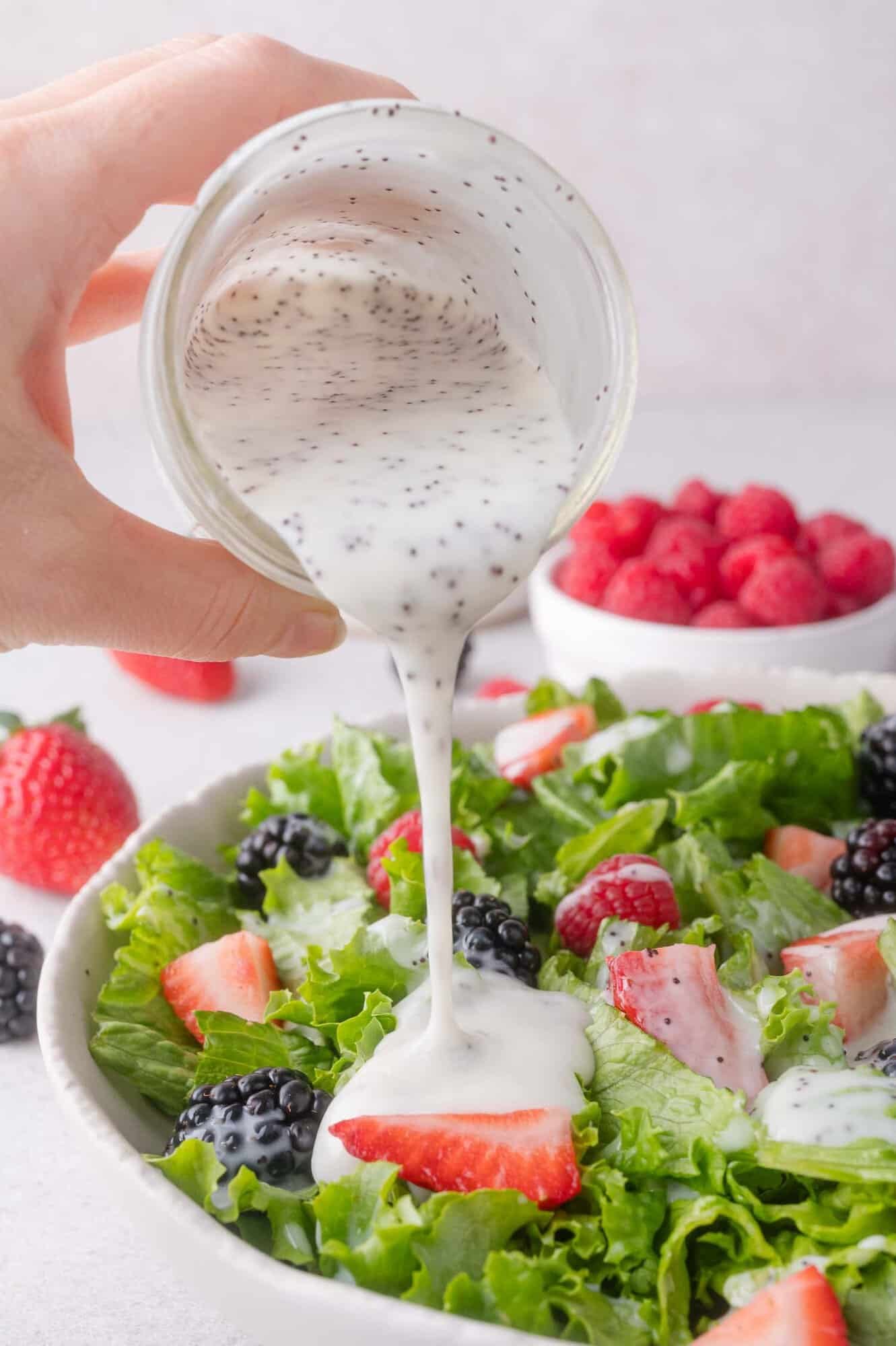 A hand pours creamy poppy seed dressing over a green salad with blackberries, strawberries, and raspberries.