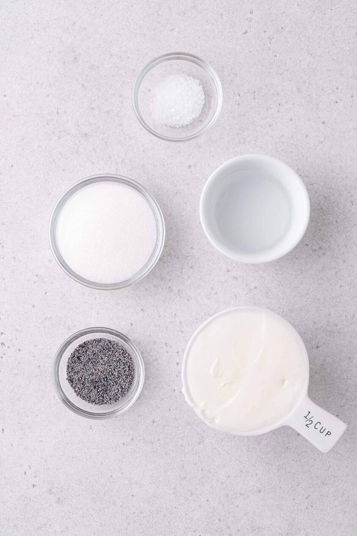Five small bowls on a light countertop containing salt, sugar, poppy seeds, white vinegar, and mayonnaise.