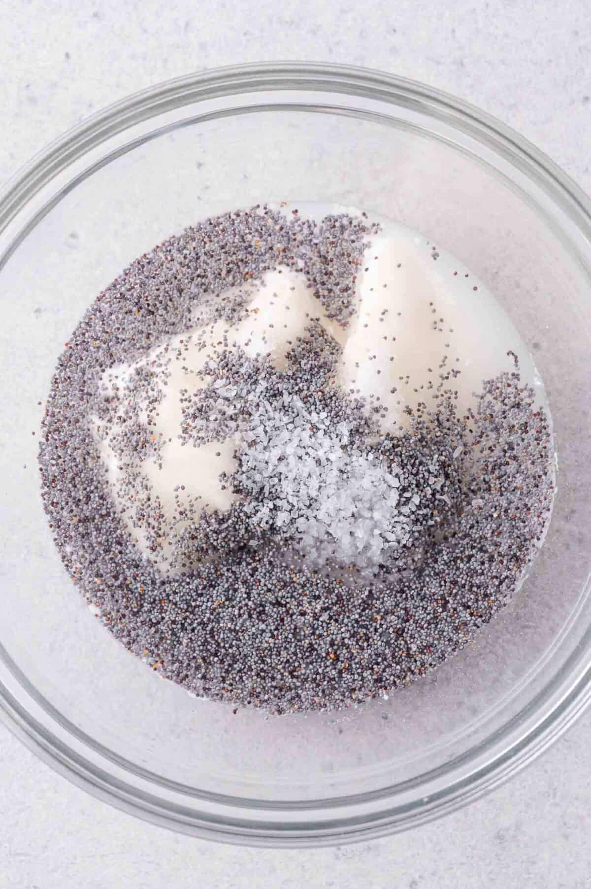A clear glass bowl containing poppy seeds, mayonnaise, sugar, and salt on a light-colored surface.