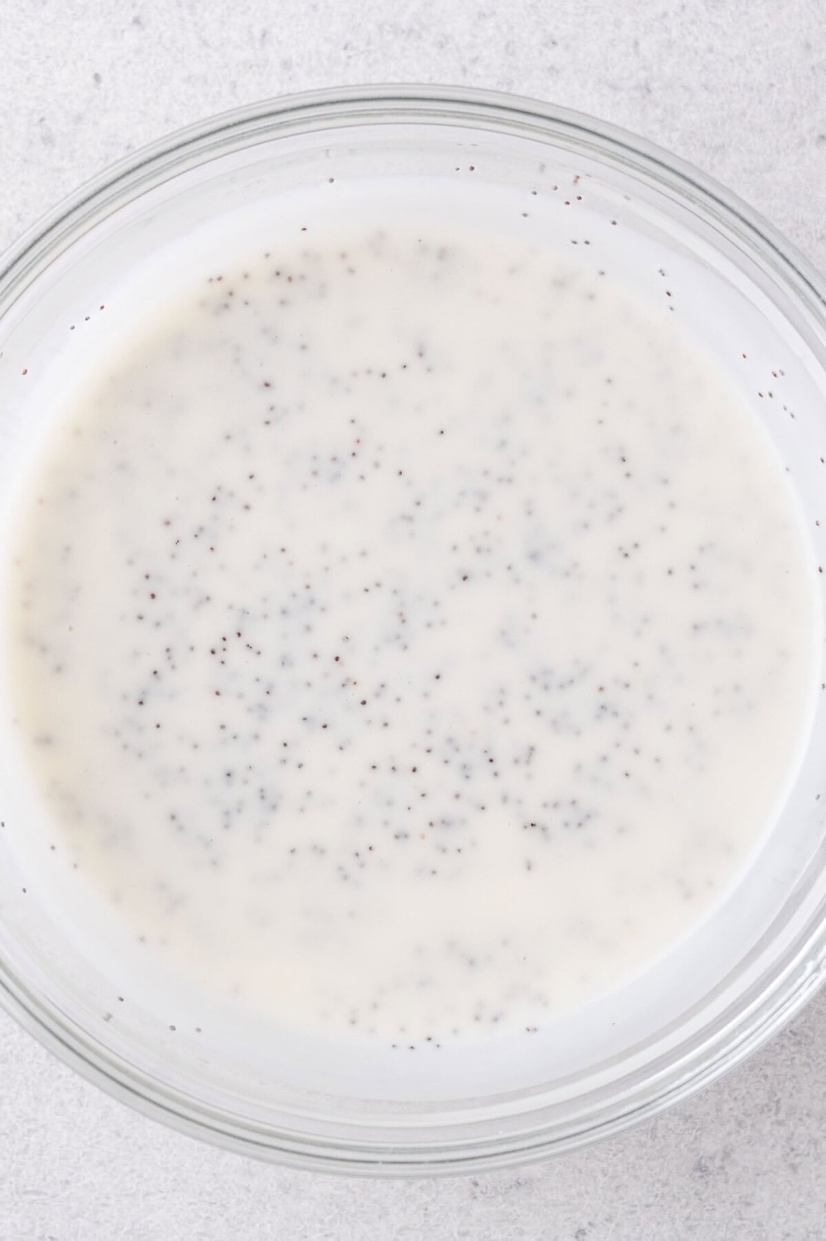 A clear glass bowl filled with a creamy white liquid containing small black poppy seeds, sitting on a light gray surface.