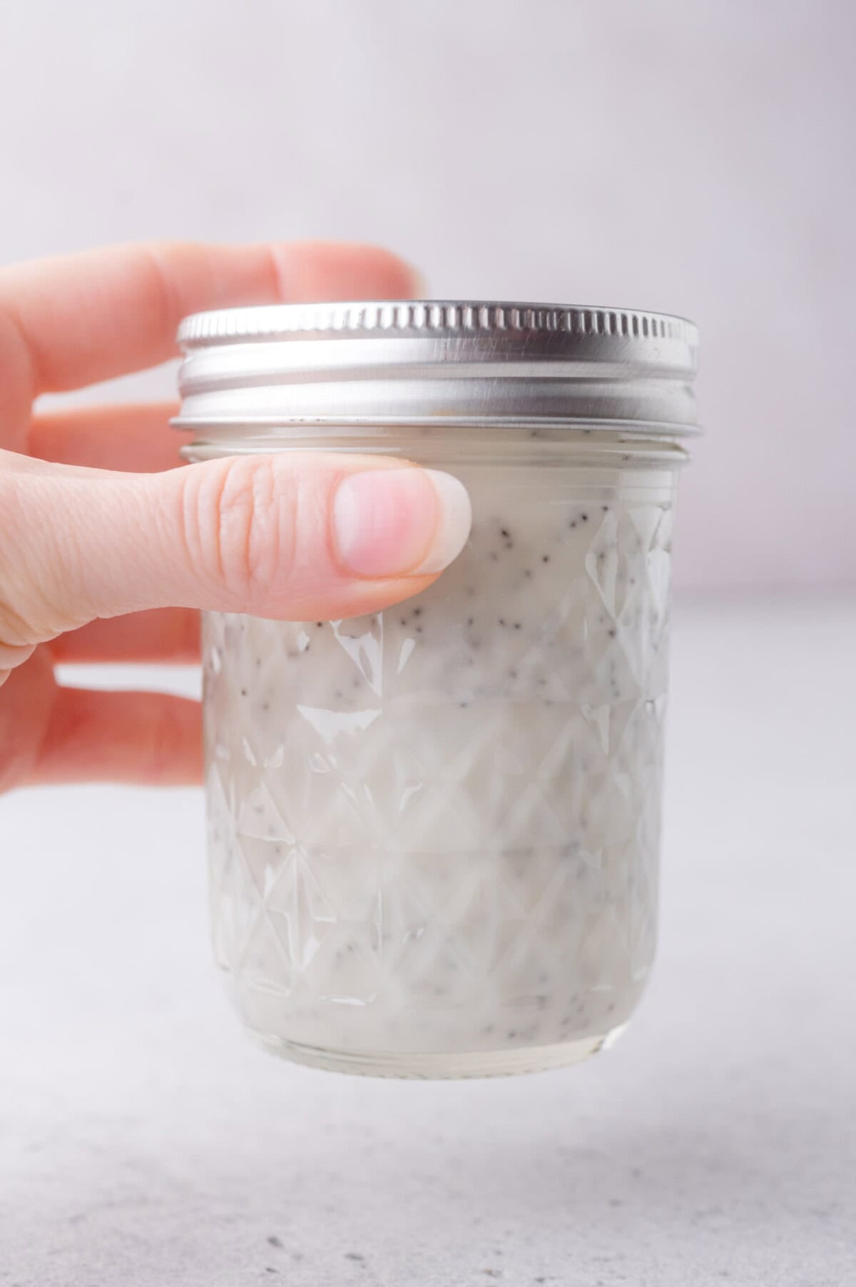 A hand holds a glass mason jar with a metal lid, filled with a creamy, poppy seed dressing.