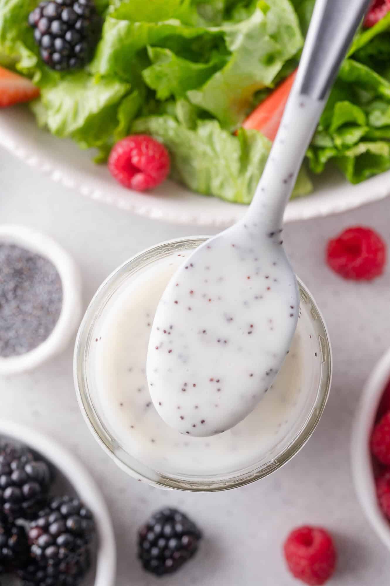 A spoonful of creamy poppy seed dressing held above a jar, surrounded by fresh berries and a bowl of salad greens.
