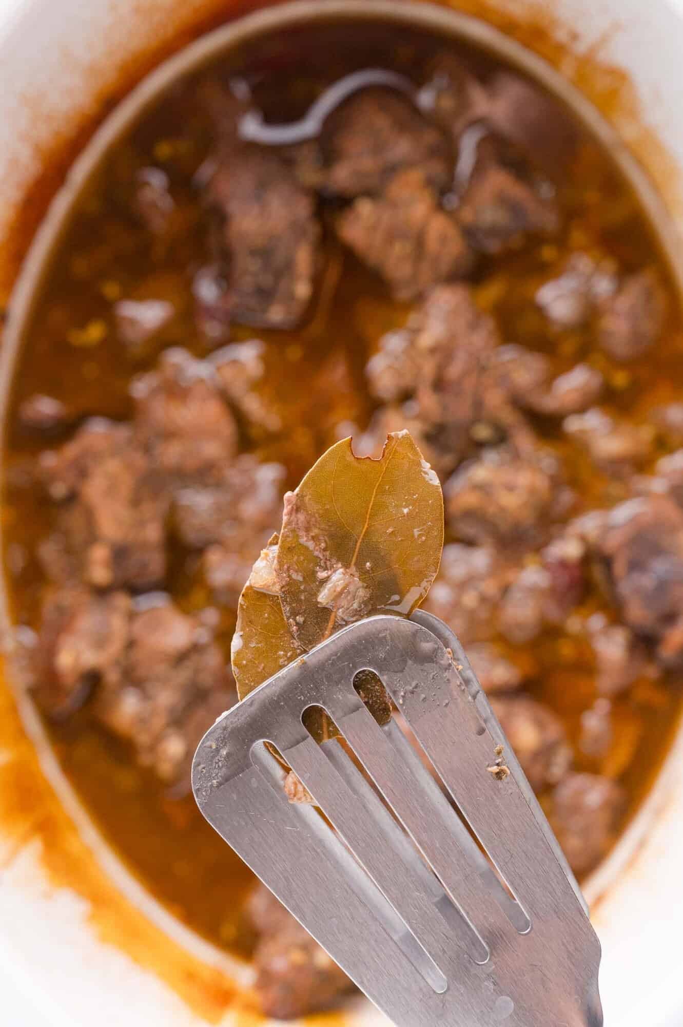 A close-up of a metal spatula holding a bay leaf above a dish of cooked beef in a chipotle sauce.