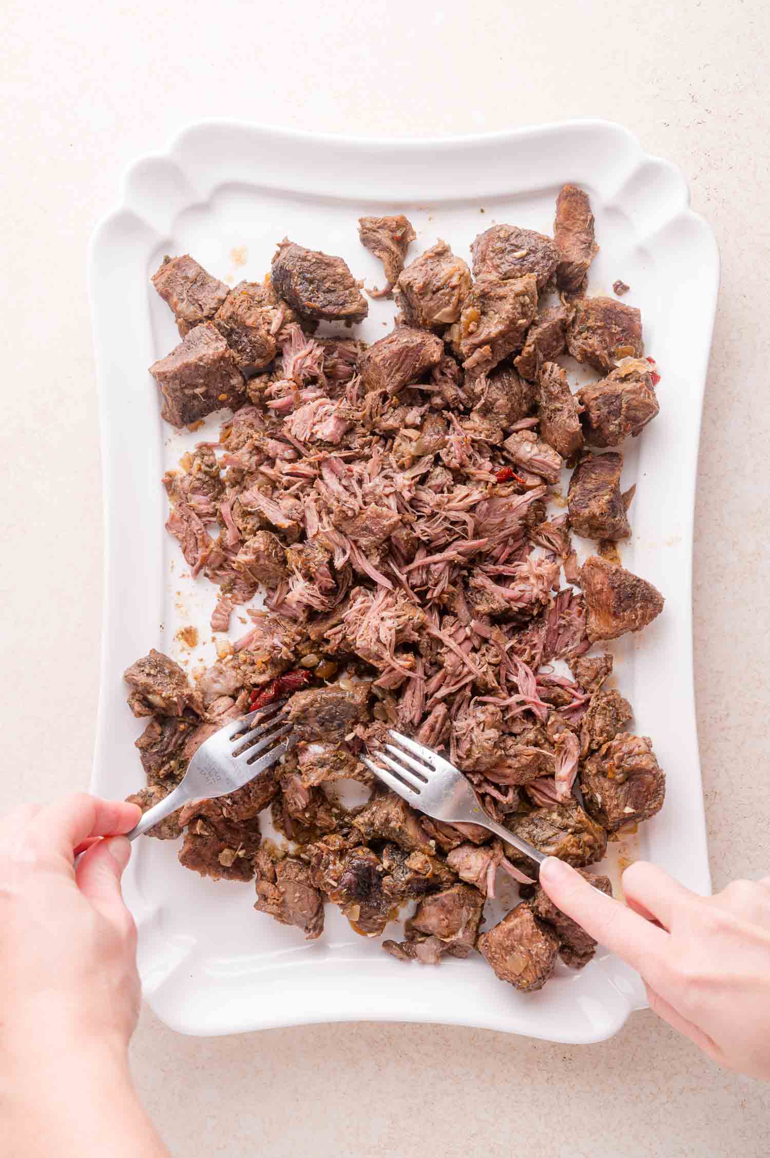 Two hands use forks to shred cooked beef chunks on a white platter.