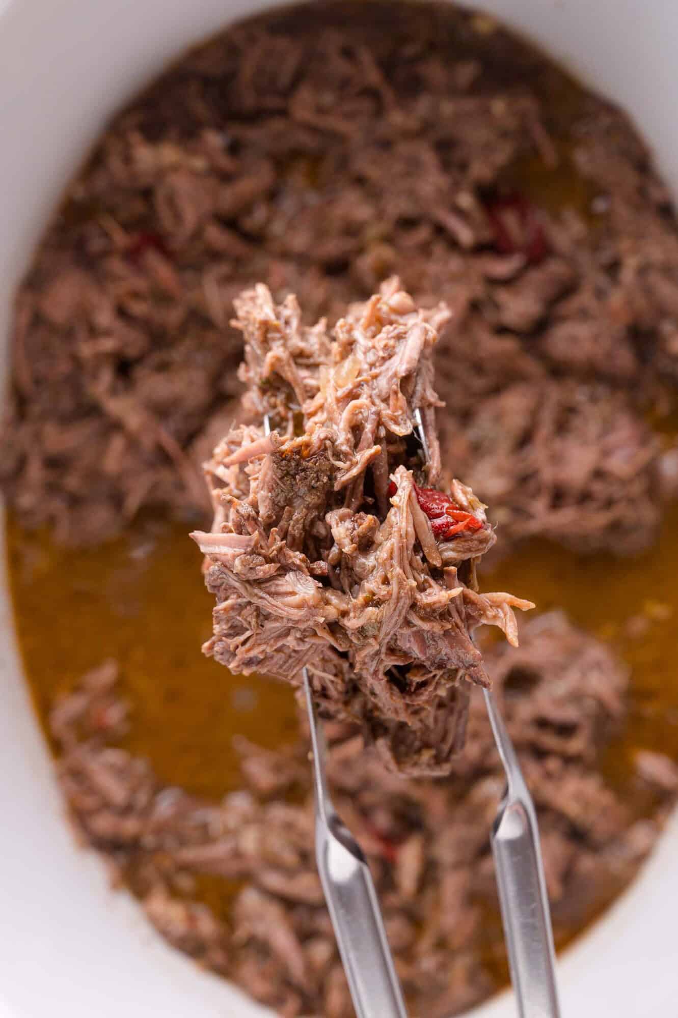 Close-up of metal tongs holding shredded beef barbacoa above a dish filled with more shredded beef and broth.