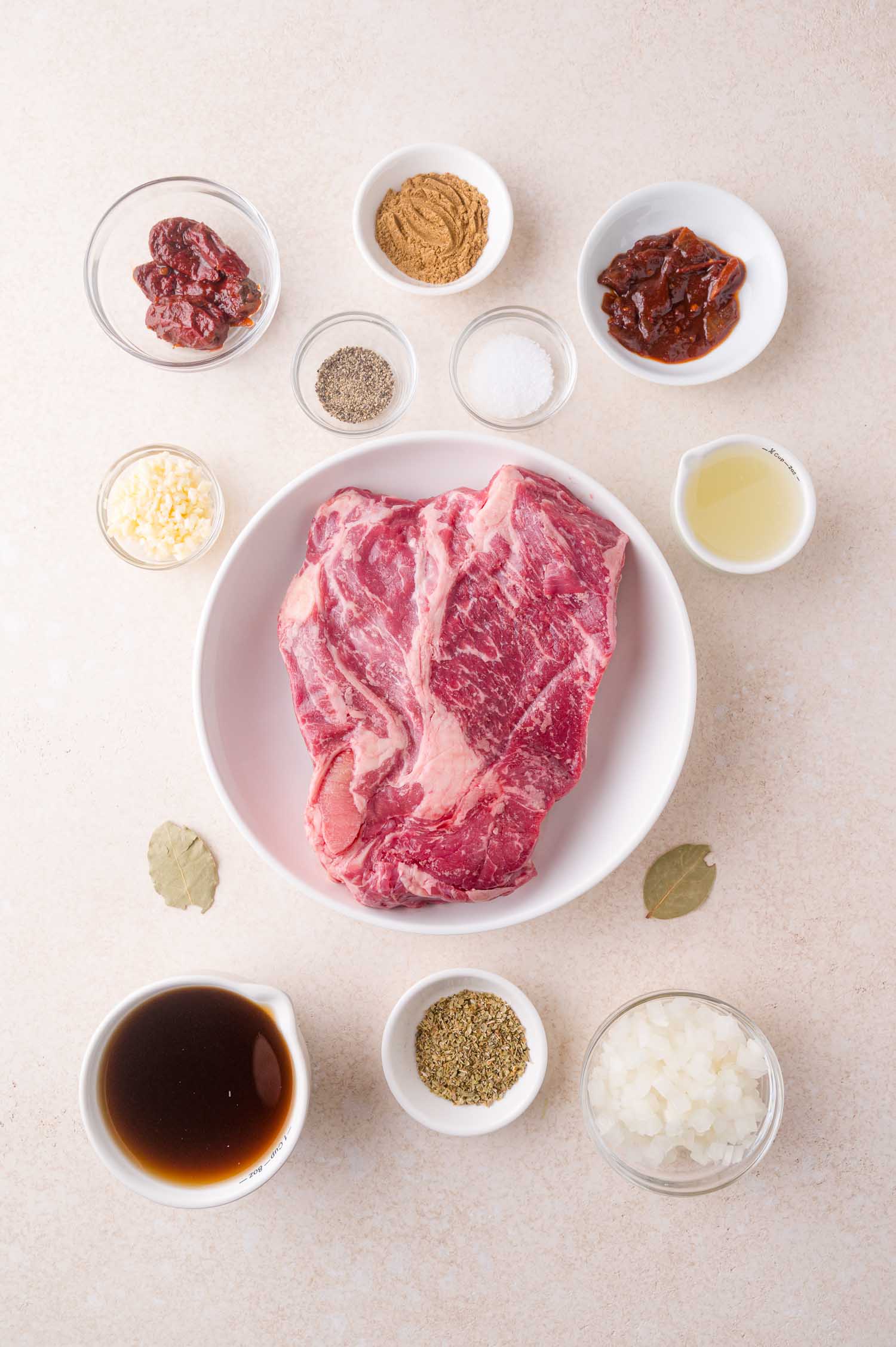 A raw marbled cut of beef in a bowl surrounded by small bowls containing various spices, chopped onion, sauce, and other ingredients on a light surface.