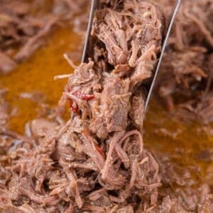 Close-up of shredded beef in a pot with visible juices and pieces of red pepper, being lifted with metal tongs.