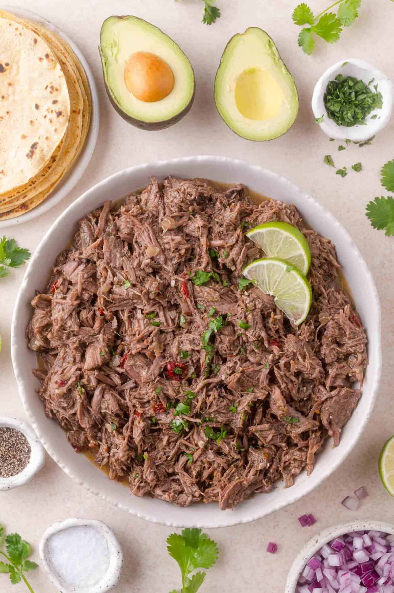 A bowl of shredded beef garnished with lime slices, surrounded by tortillas, avocado halves, chopped herbs, red onion, and seasoning bowls on a light surface.