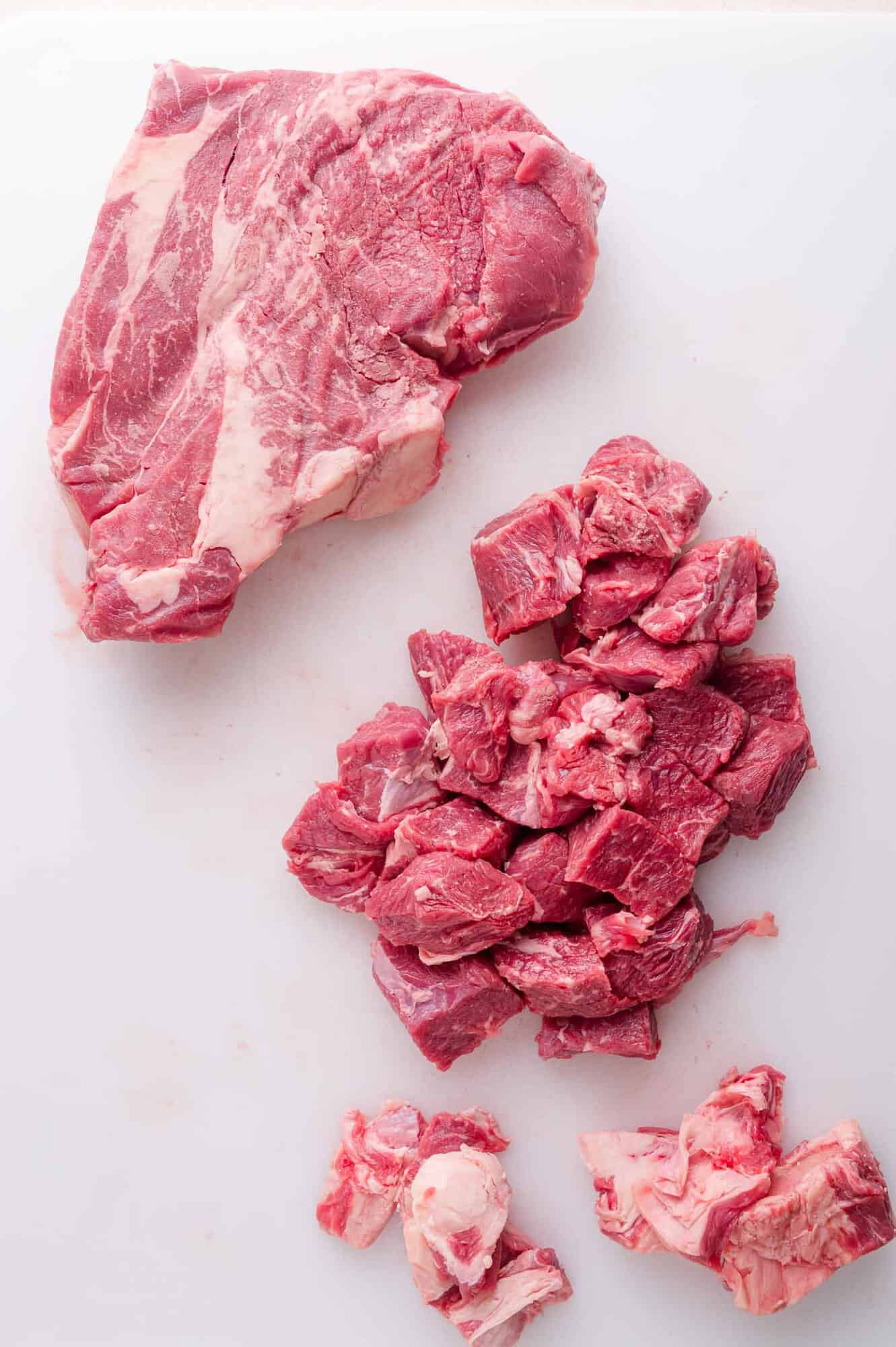 A large piece of raw beef, diced chunks of beef, and small beef trimmings arranged on a white cutting board.