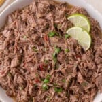 Shredded beef barbacoa in a white bowl, garnished with chopped cilantro and two lime slices.