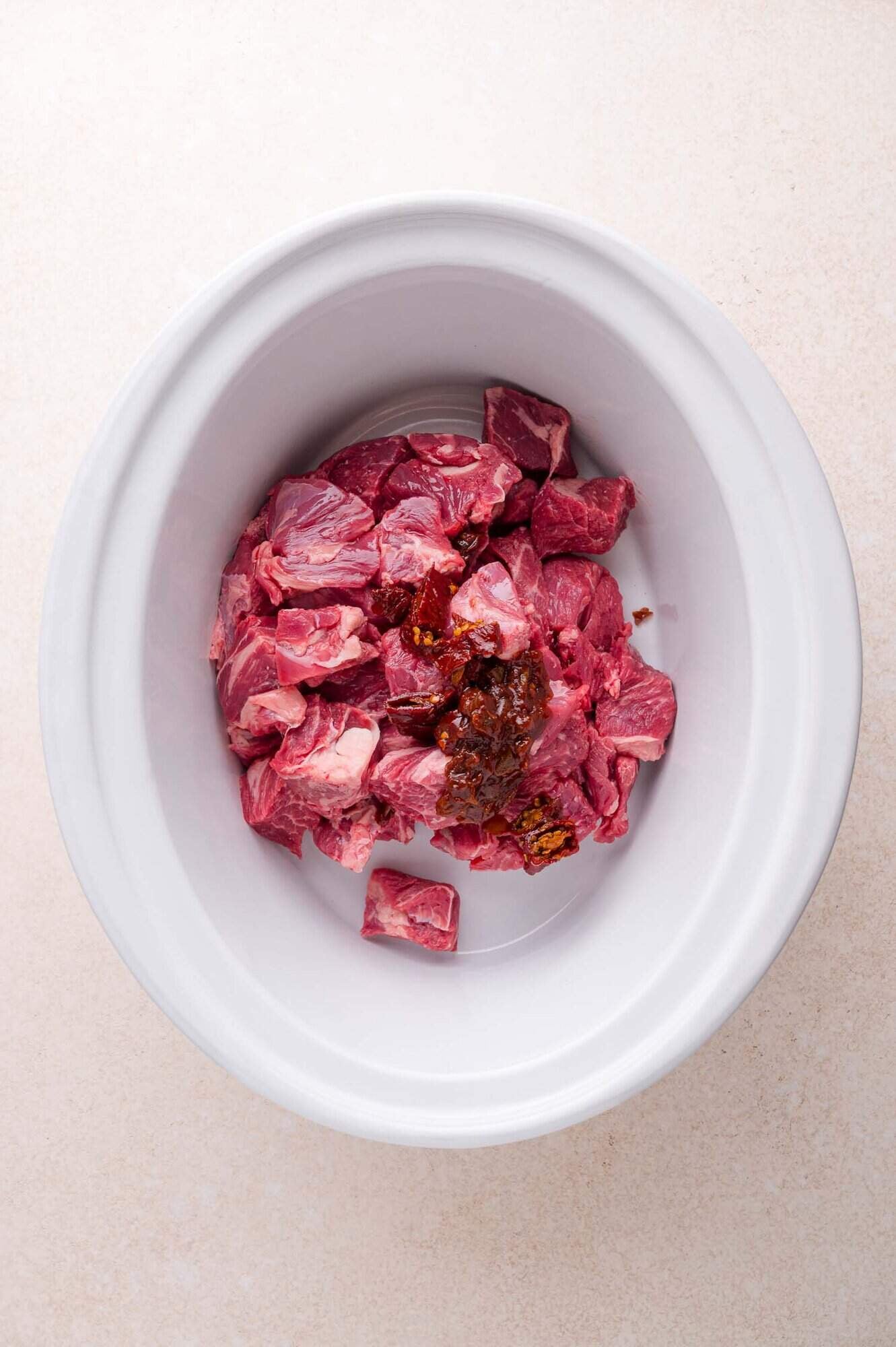 Raw beef chunks and a dollop of chipotle peppers are placed in a white slow cooker on a light-colored surface.