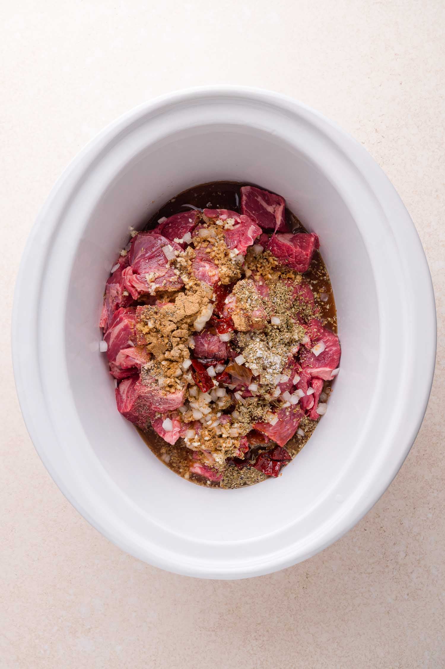 Cubed raw beef with various spices in a white slow cooker, ready to be cooked.