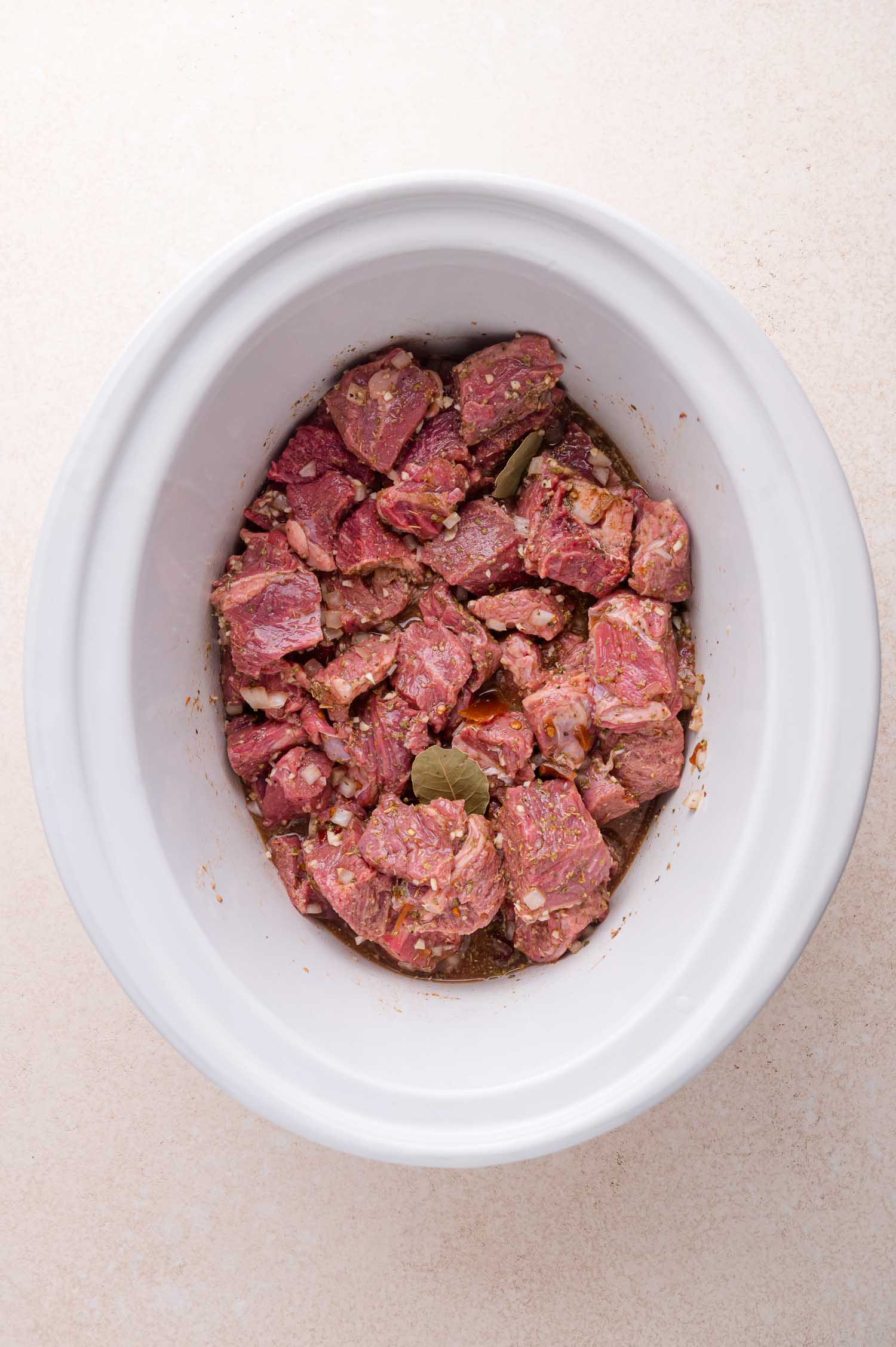 Chunks of raw, marinated beef with spices and bay leaves placed in a white slow cooker, ready to be cooked.
