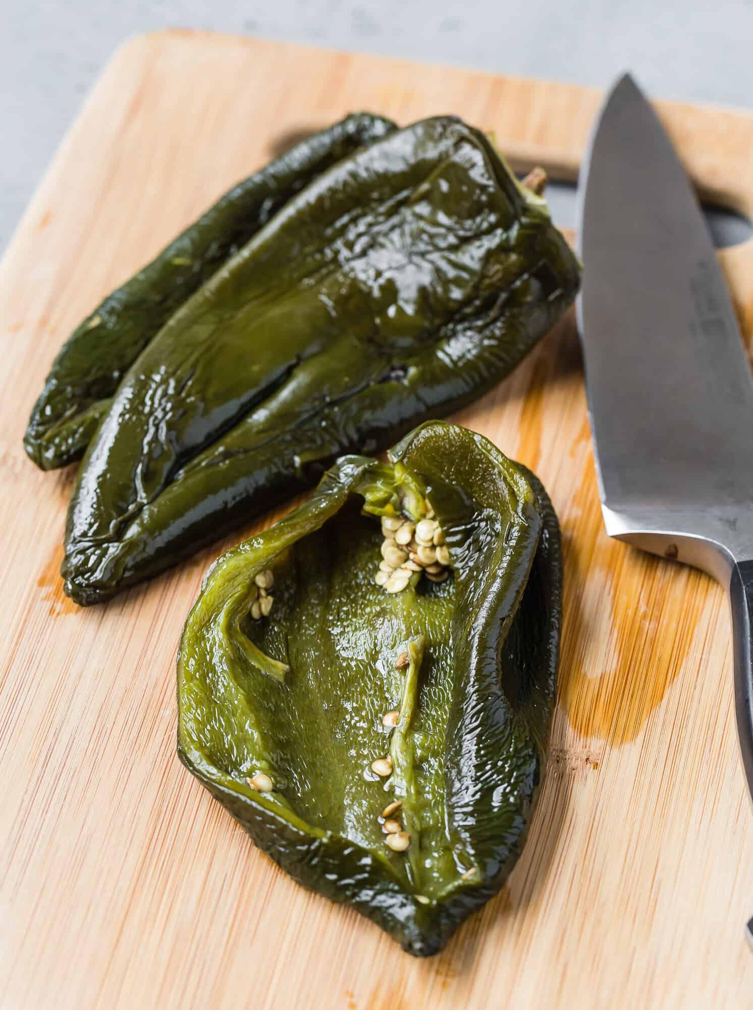 Two roasted poblano peppers, one sliced open to show seeds inside, on a wooden cutting board next to a kitchen knife.
