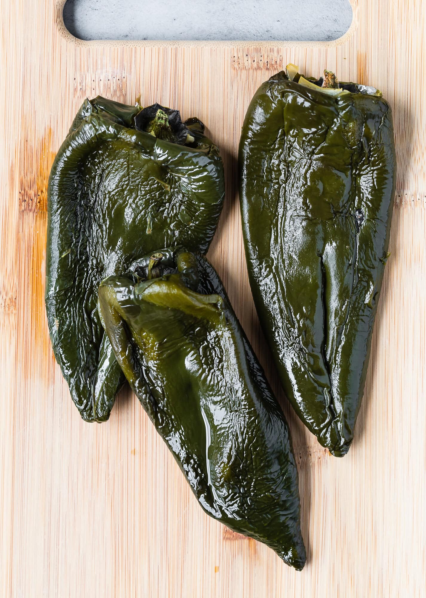 Three roasted poblano peppers on a wooden cutting board.