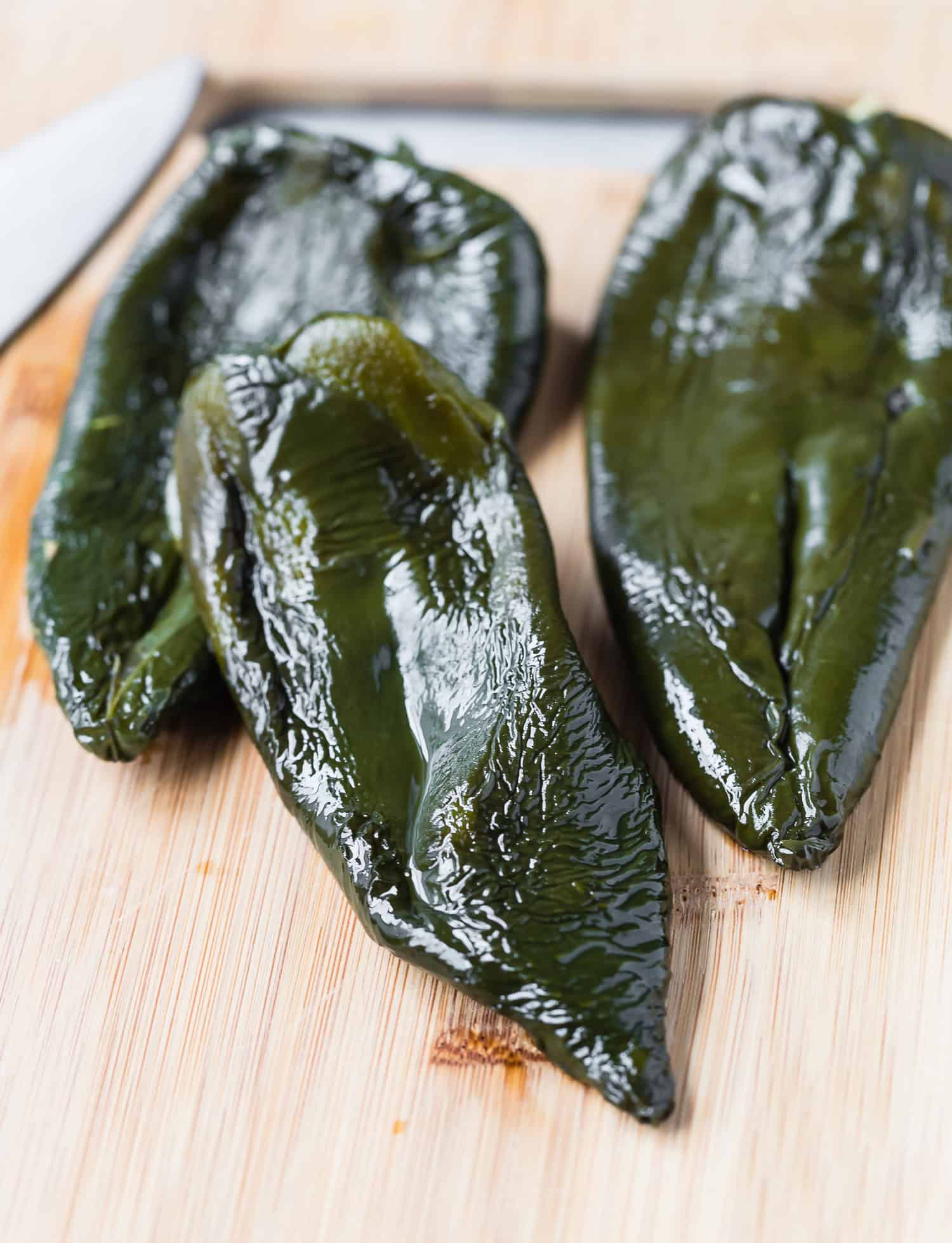 Three roasted poblano peppers on a wooden cutting board with a knife in the background.