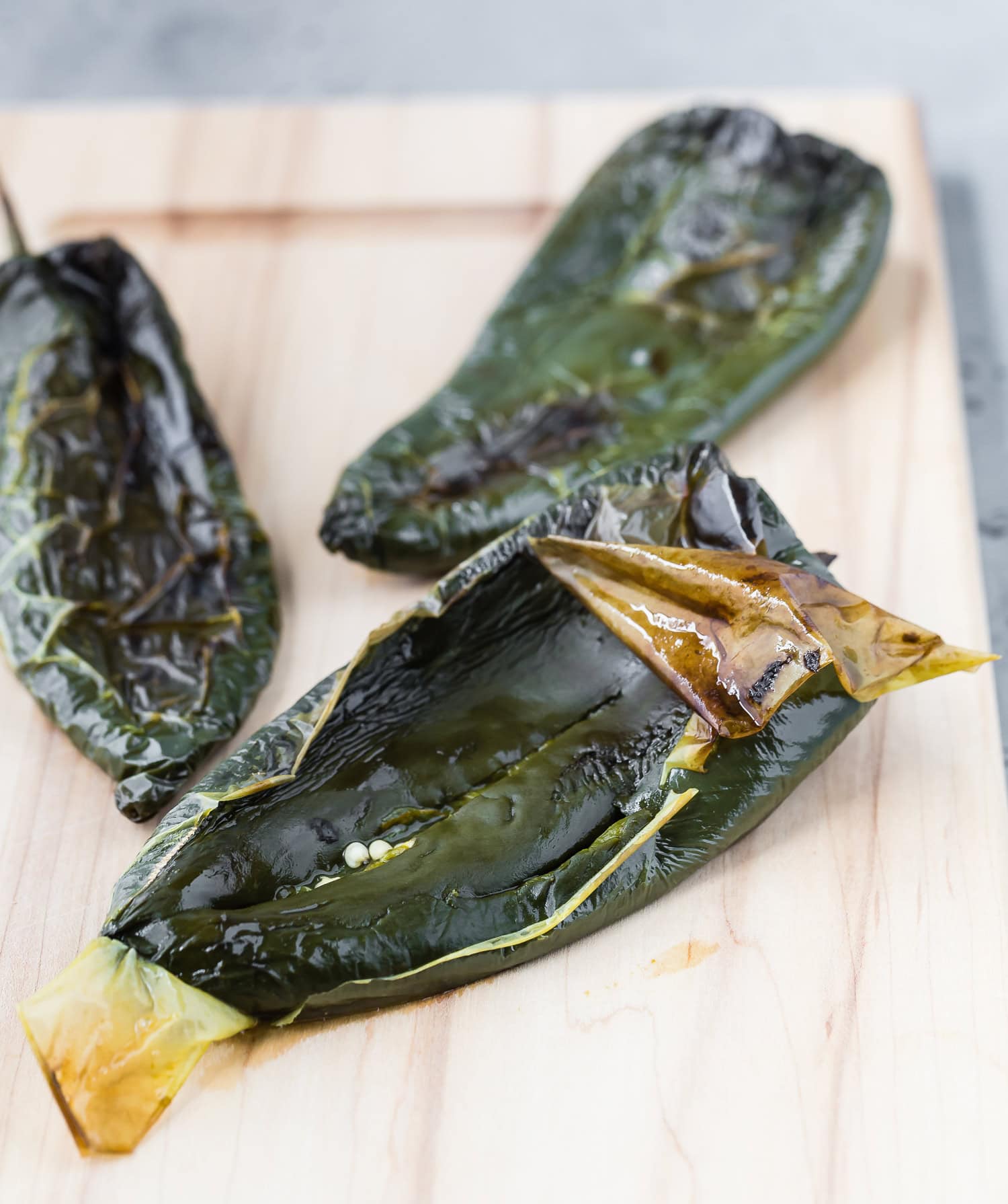 Three charred poblano peppers with blistered skins rest on a light wooden surface.