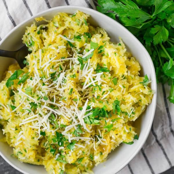 A bowl of cooked spaghetti squash topped with grated cheese and chopped parsley, placed on a striped cloth with fresh parsley on the side.