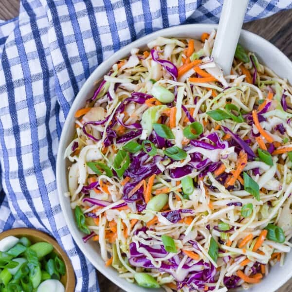 A bowl of coleslaw with shredded cabbage, carrots, and green onions sits on a blue checkered cloth, with a small bowl of chopped green onions and salt and pepper shakers nearby.