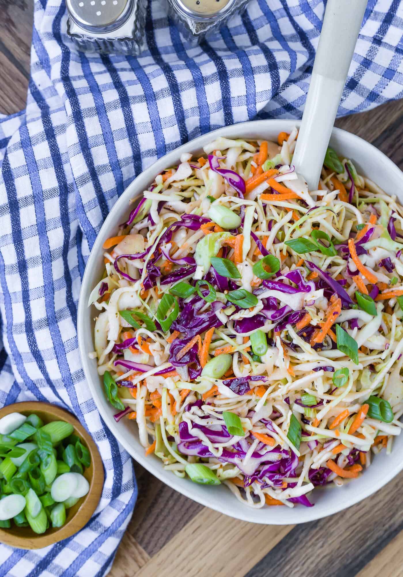 A bowl of coleslaw with shredded cabbage, carrots, and green onions sits on a blue checkered cloth, with a small bowl of chopped green onions and salt and pepper shakers nearby.