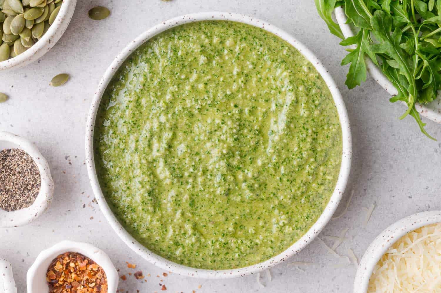A bowl of arugula pesto sauce surrounded by bowls of arugula, grated cheese, red pepper flakes, black pepper, and pumpkin seeds.
