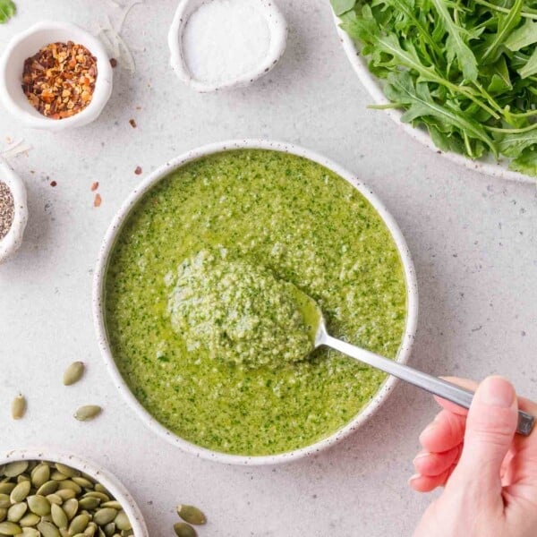 A hand holds a spoon scooping green arugula pesto from a bowl, surrounded by arugula, pumpkin seeds, red pepper flakes, salt, and pepper.