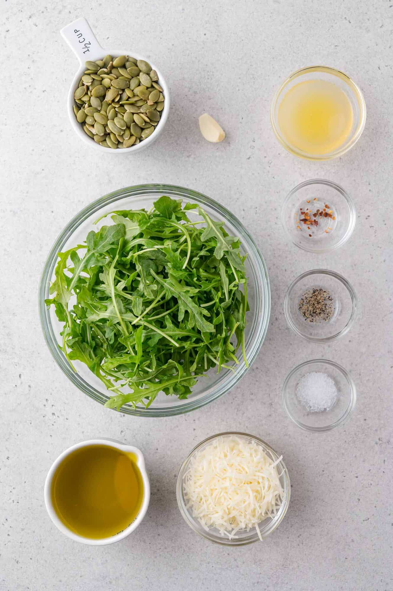 A bowl of arugula surrounded by small bowls of pumpkin seeds, olive oil, grated cheese, lemon juice, a garlic clove, salt, pepper, and red pepper flakes on a light surface.