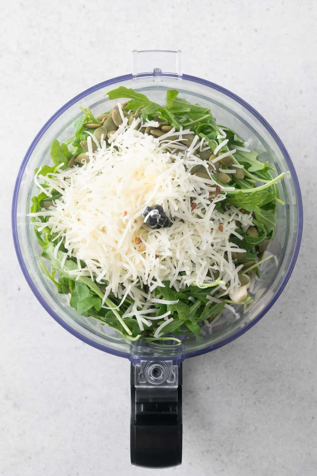 A food processor bowl filled with arugula leaves, grated cheese, pepitas, and seasonings, viewed from above.