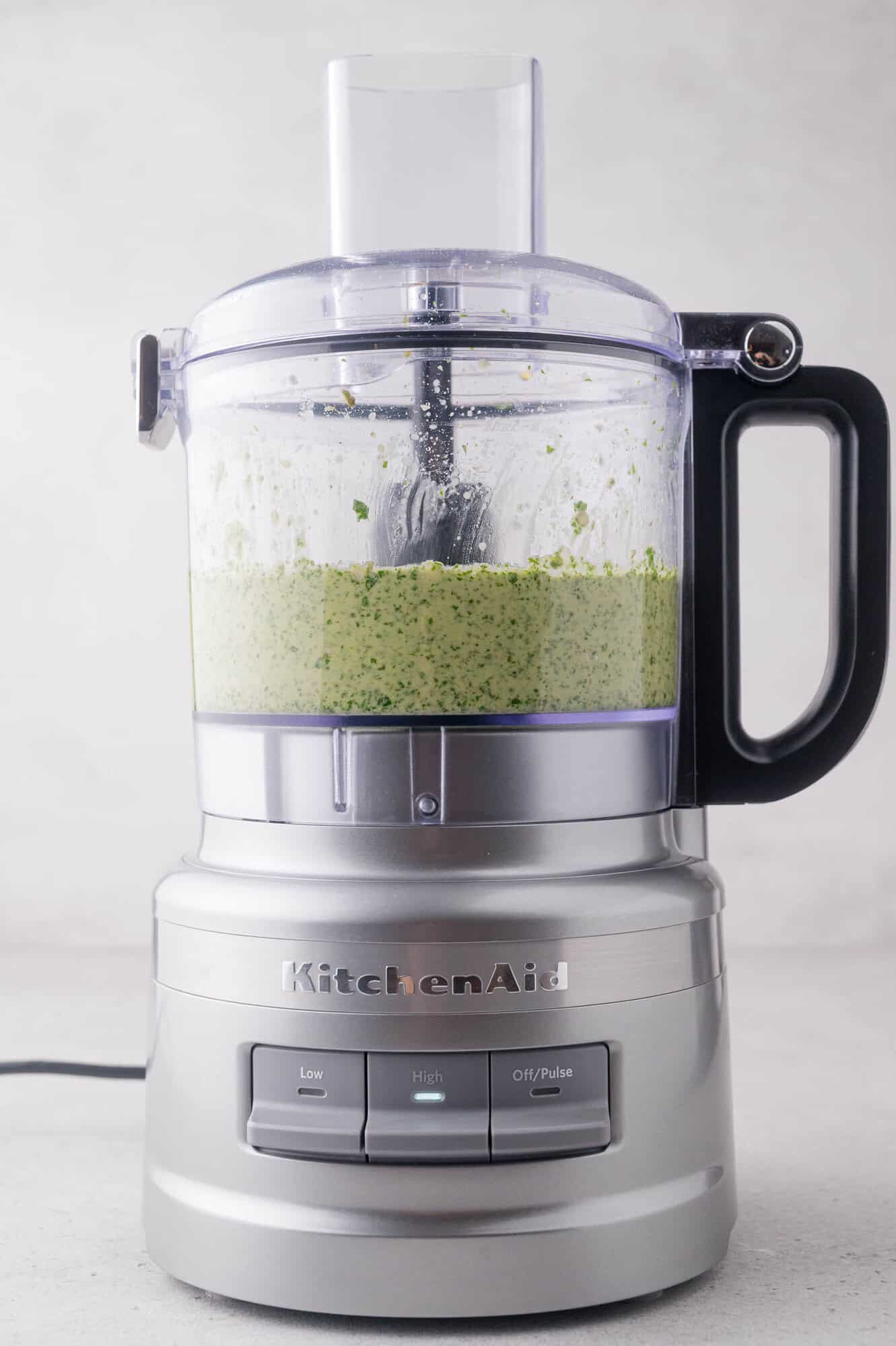 A silver KitchenAid food processor blending arugula pesto, placed on a light gray surface.