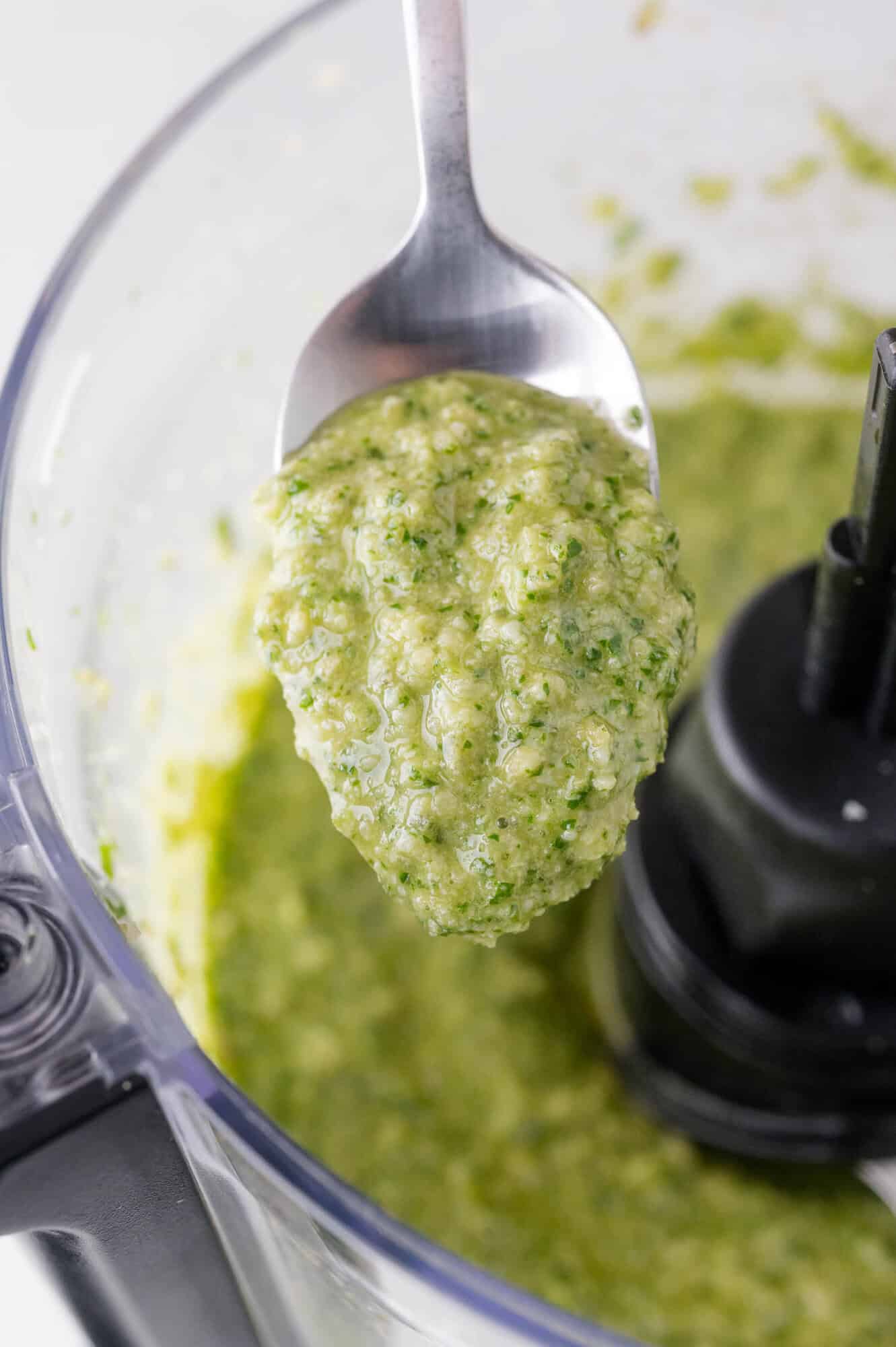 A spoon holds a thick green arugula pesto above a food processor filled with more of the same blended substance.