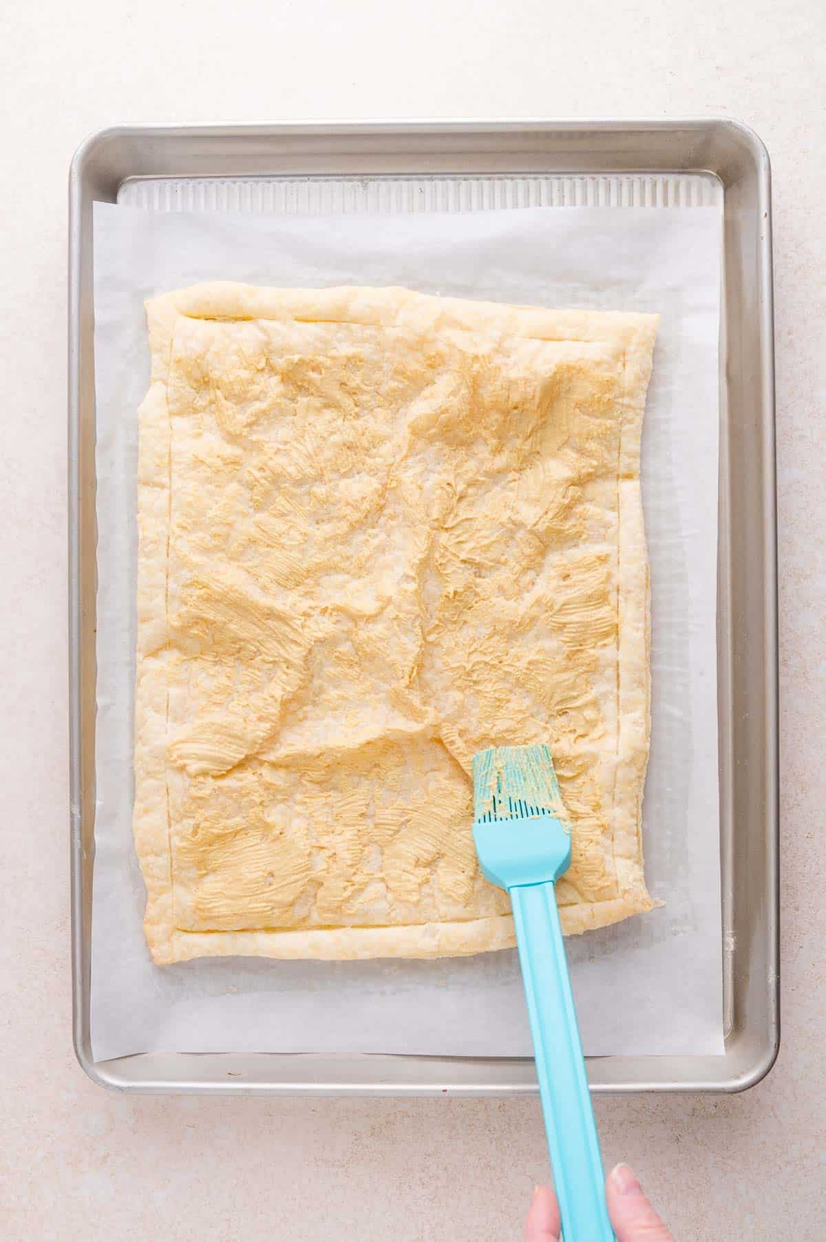 A hand uses a blue silicone brush to apply an egg wash to a sheet of puff pastry on a parchment-lined baking tray.