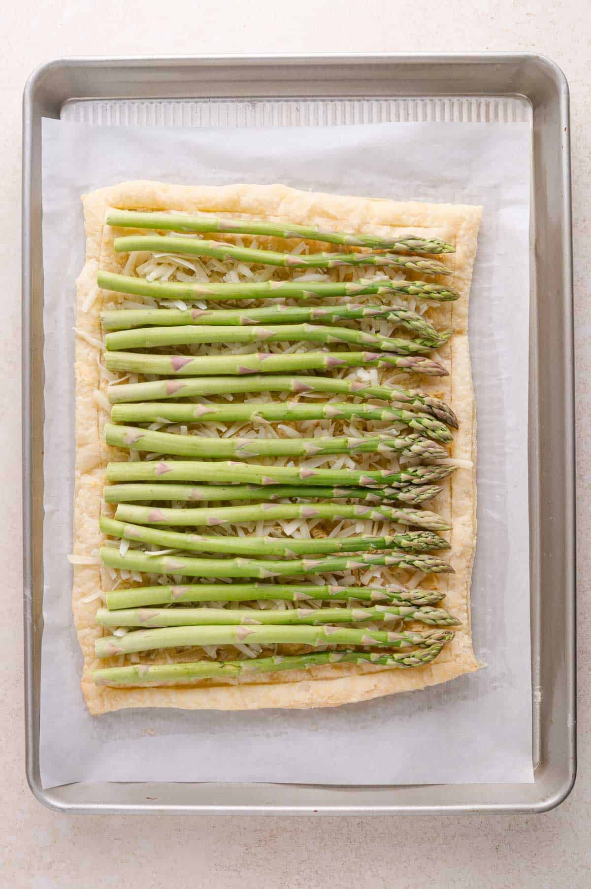 Rectangular puff pastry topped with shredded cheese, and raw asparagus spears, arranged on a baking sheet lined with parchment paper.