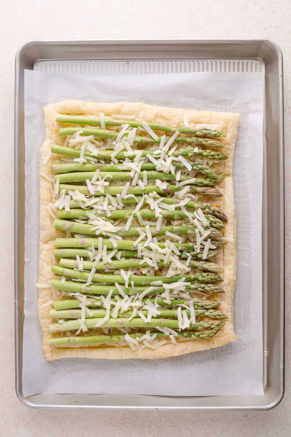 Rectangular puff pastry topped with asparagus spears and shredded cheese, placed on parchment paper in a baking tray.