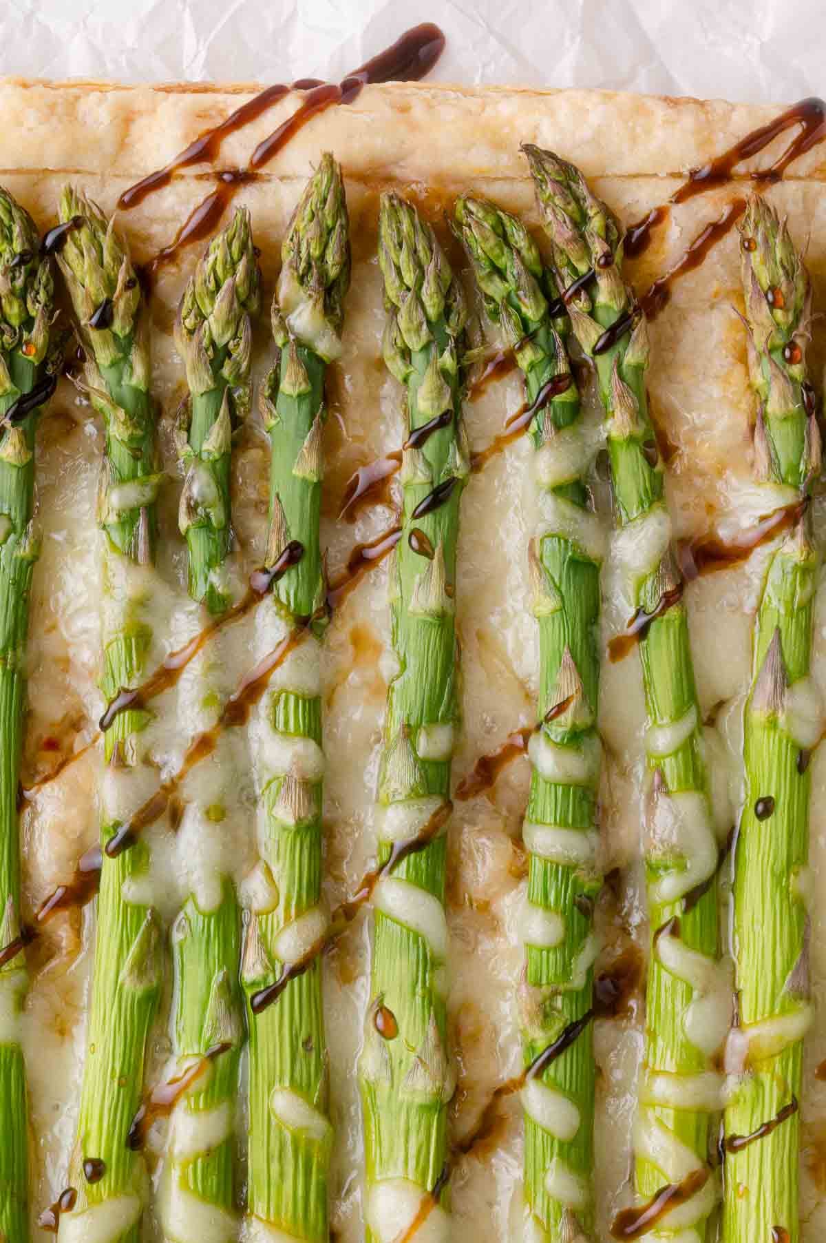 Close-up of asparagus spears arranged on a puff pastry topped with melted cheese and drizzled with balsamic glaze.