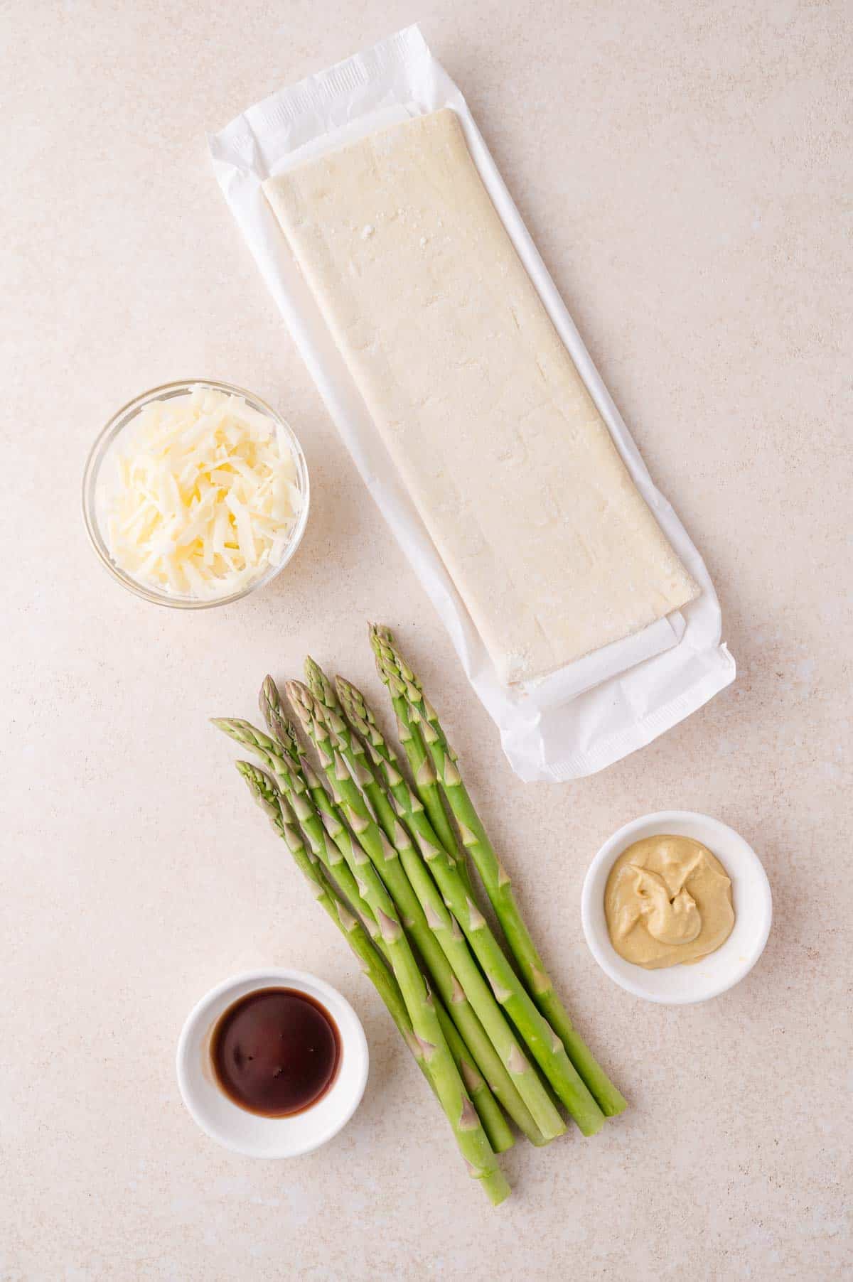 Five ingredients on a light surface: a block of puff pastry, shredded cheese in a bowl, fresh asparagus spears, and small bowls of mustard and sauce.