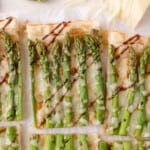 Rectangular slices of puff pastry topped with asparagus, melted cheese, and a drizzle of balsamic glaze, arranged on parchment paper.