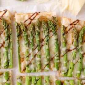 Rectangular slices of puff pastry topped with asparagus, melted cheese, and a drizzle of balsamic glaze, arranged on parchment paper.