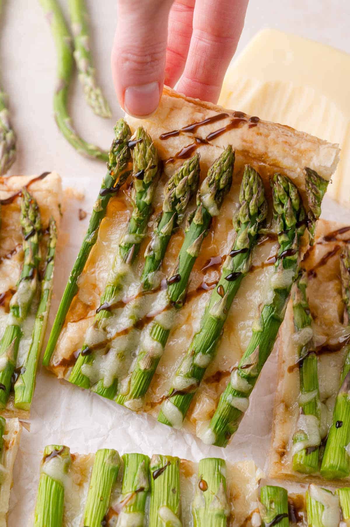 A hand holding a rectangular slice of puff pastry topped with melted cheese, asparagus spears, and a drizzle of balsamic glaze.
