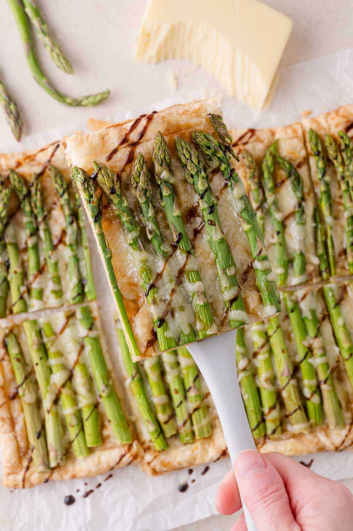 A hand holds a spatula lifting a slice of asparagus tart with melted cheese and balsamic drizzle from a parchment-lined surface.
