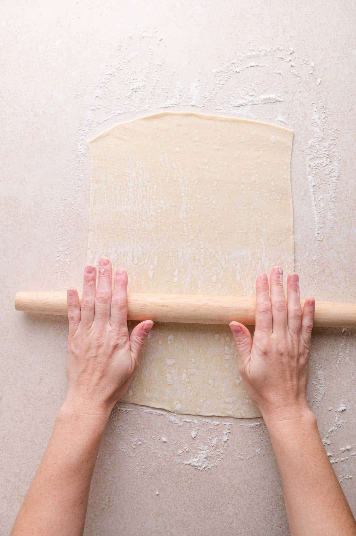 Hands using a rolling pin to flatten puff pastry on a lightly floured surface.