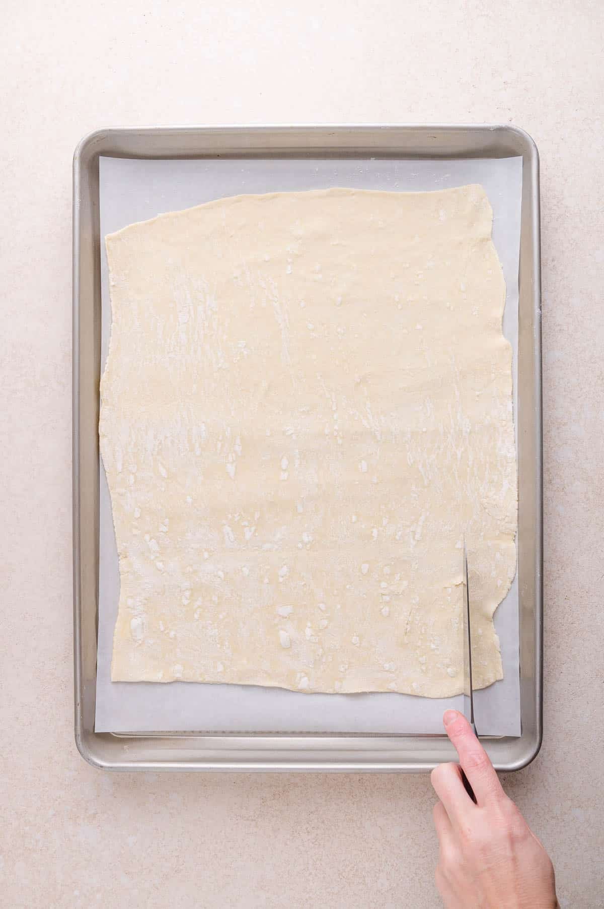 A hand uses a knife to cut a large sheet of parchment paper placed on a parchment-lined baking tray.