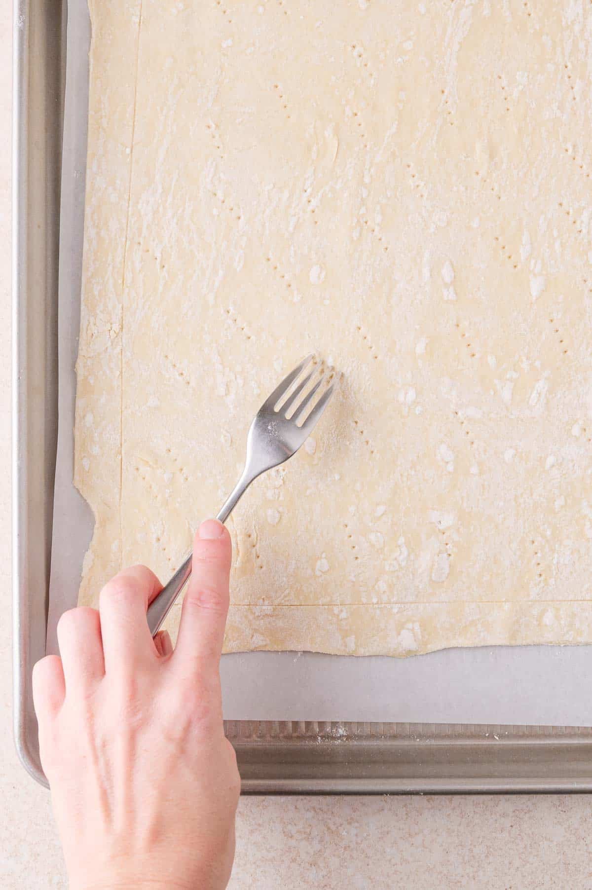 A hand presses a fork into a sheet of raw puff pastry dough placed on a baking tray lined with parchment paper.