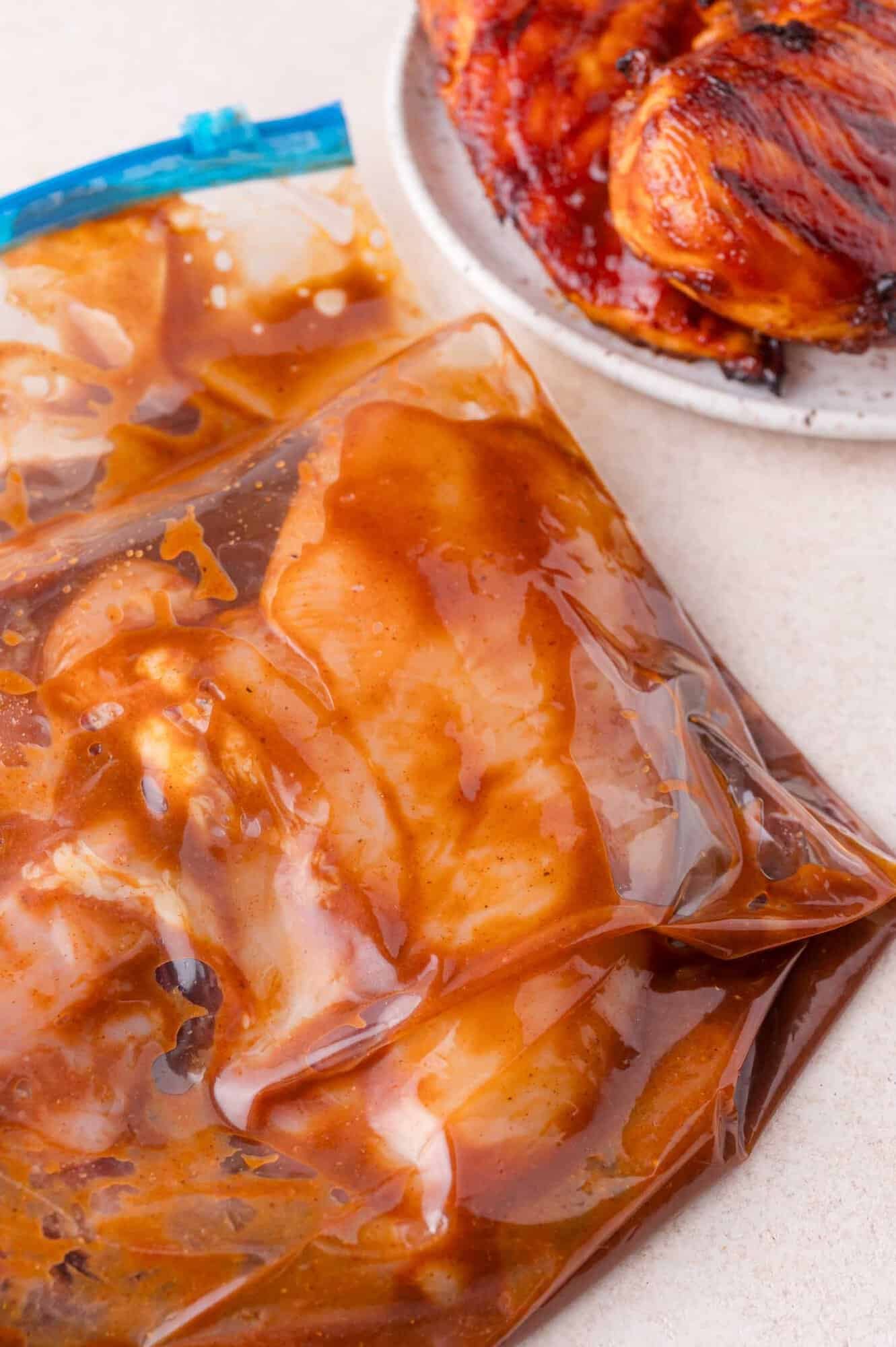 Raw chicken breasts marinating in a sealed plastic bag with sauce, next to a plate of cooked, glazed chicken.