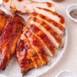 Sliced grilled chicken breast coated in barbecue sauce on a white plate, with small bowls of pepper and salt nearby.