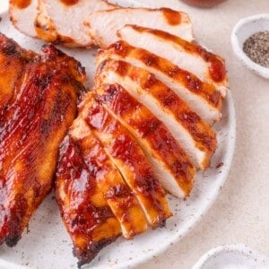 Sliced grilled chicken breast coated in barbecue sauce on a white plate, with small bowls of pepper and salt nearby.