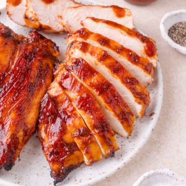Sliced grilled chicken breast coated in barbecue sauce on a white plate, with small bowls of pepper and salt nearby.