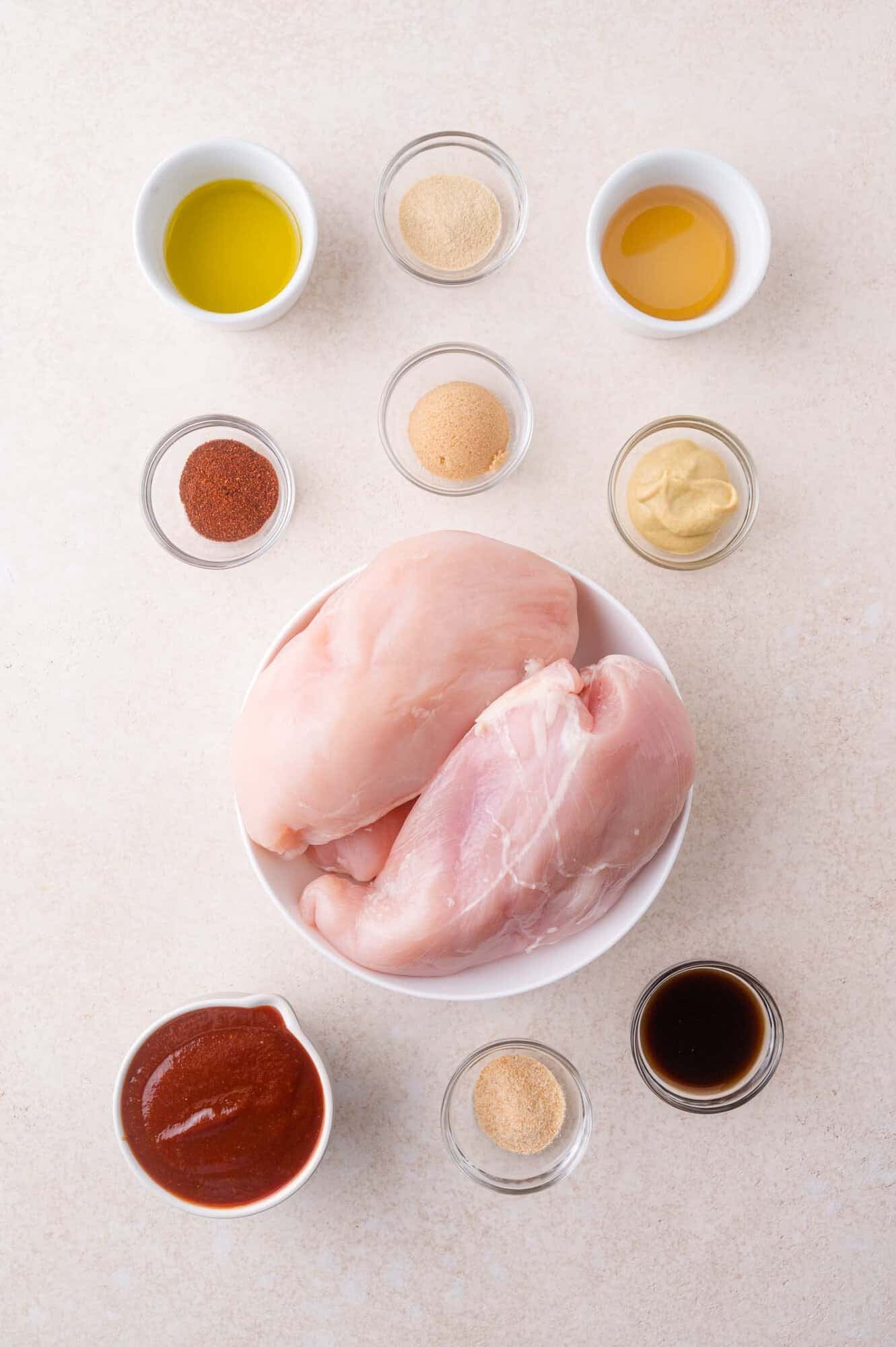 Raw chicken breasts in a bowl surrounded by small bowls containing various spices, sauces, and seasonings arranged on a light surface.