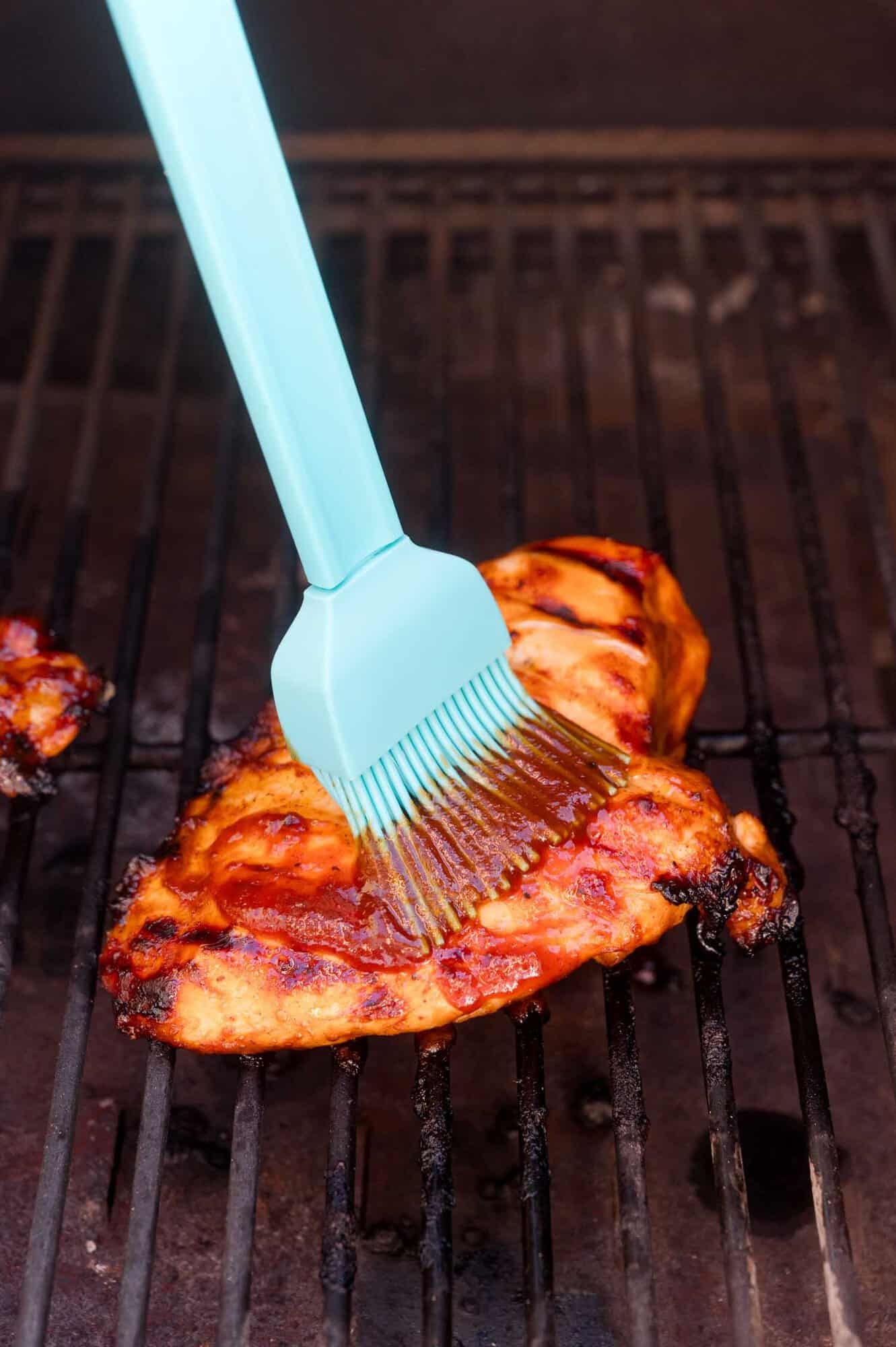 A blue silicone brush applies sauce to a grilled chicken breast on a barbecue grill.