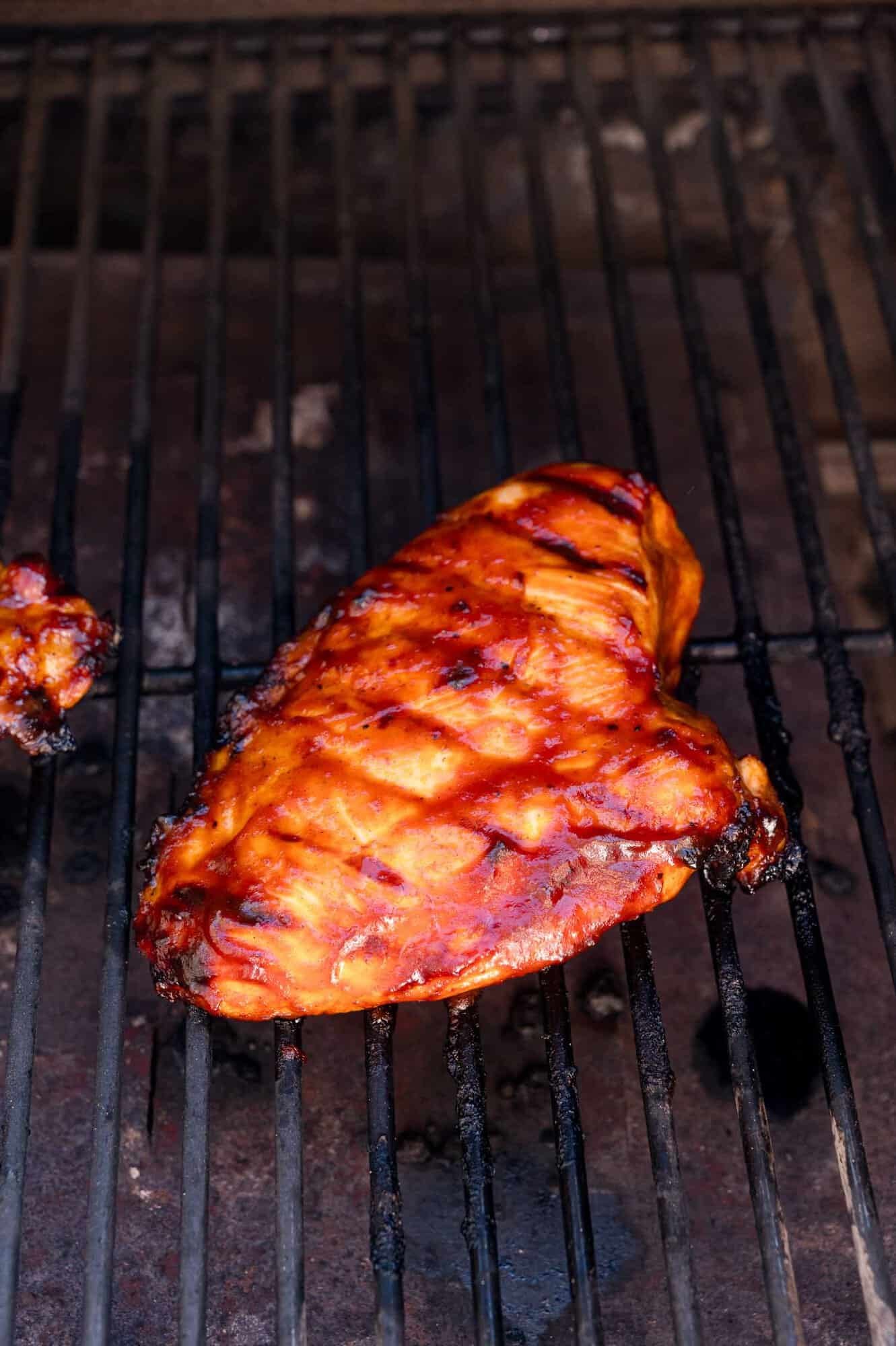 A grilled chicken breast coated in barbecue sauce sits on a hot grill with visible grill marks.